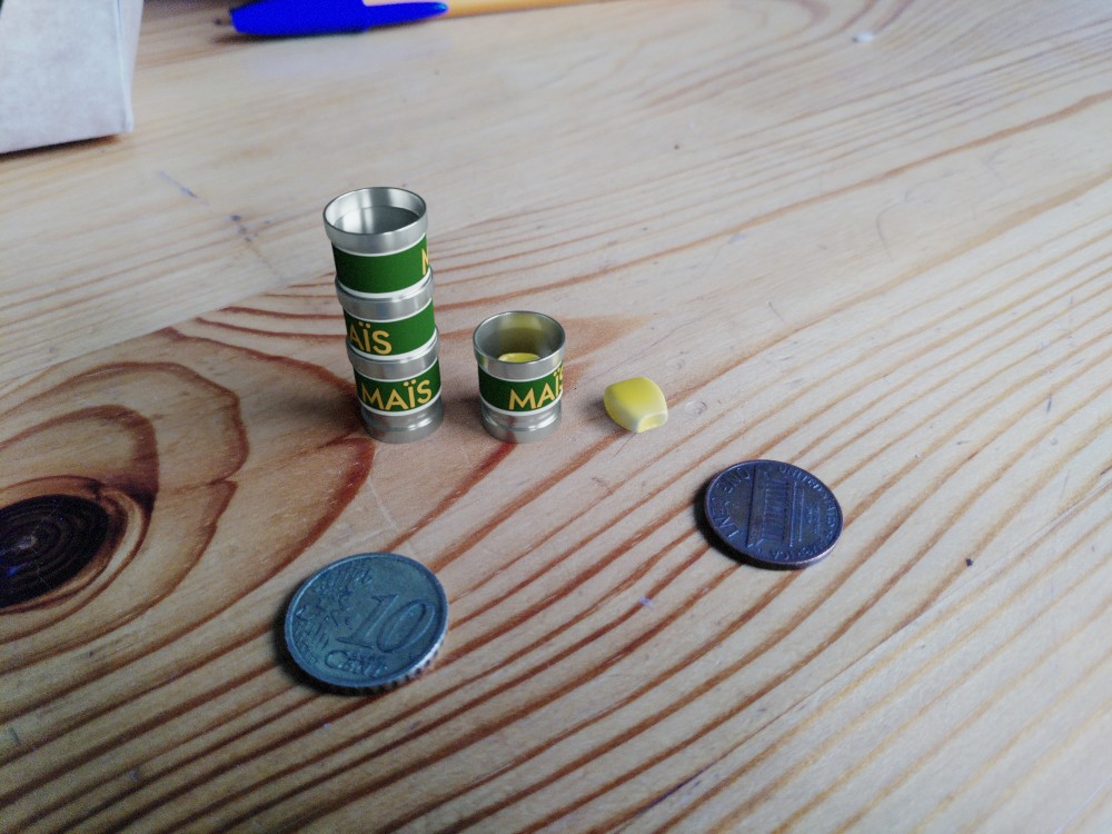 A composite image featuring a wooden table, atop of which are tins of maize, one centimetre in diameter and containing <em>one</em> grain. Two coins, one of ten euro cents, one of one dollar cent, are set next to the tins for scale.